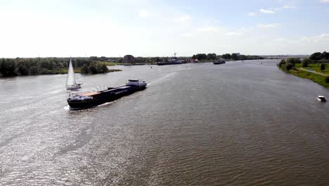 Aerial-Over-Oude-Maas-With-Draga-Cargo-Ship-Going-Past-Sailing-Boat-On-Sunny-Day