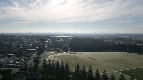 Aerial-flyover-a-neighborhood-residential-housing,-park-and-sports-field-with-Sydney-Skyline-in-silhouette