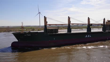 Aerial-Port-Side-View-Of-Aal-Paris-Cargo-Ship-Travelling-Along-Oude-Maas-With-Still-Wind-Turbine-In-Background
