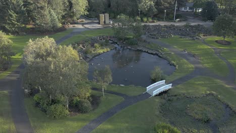 Botanical-garden-with-duck-and-geese-filled-pond,-shadow-from-trees-by-sunset,-aerial