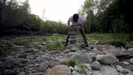 Frustrated-Male-Hiker-balances-large-pile-of-jagged-stones-with-scattered-rocks-and-dark-forest-background