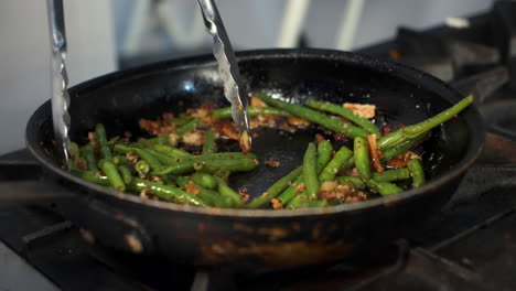 Chef-uses-tongs-to-toss-and-stir-sauteed-green-beans-in-pan,-slow-motion-close-up-4K