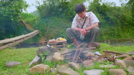 Asian-young-man-preparing-bonfire-for-a-barbecue-in-middle-of-river-meadow
