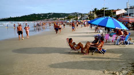 Dolly-in-of-people-relaxing-in-the-sand-under-umbrellas-near-de-shore-in-Bombas-and-Bombinhas-beaches,-Brazil