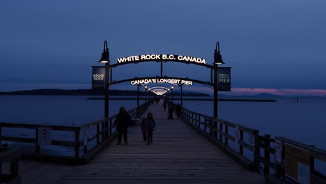 A-front-facing-view-of-the-sign-of-White-Rock-BC-Canada-World’s-longest-pier-at-night-with-people-walking-around-in-the-dark-evening-sun-setting-far-in-the-background