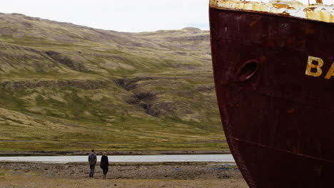 Couple-exploring-abandoned-ship-in-Iceland-coastline,-static-zoom-in-view