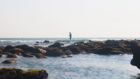 Lonely-fisherman-fishing-on-rocky-tropical-coastline,-small-ocean-waves