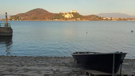 Schöne-Aufnahme-Eines-Fischerbootes,-Das-Am-Kai-Des-Lago-Maggiore-Geparkt-Ist,-Mit-Der-Festung-Von-Angera-Im-Hintergrund