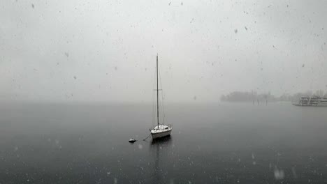 Amazing-zoom-in-shot-of-snow-falling-over-Maggiore-lake-and-moored-sailing-boat,-Italy