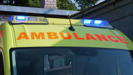 The-roof-of-yellow-paramedic-ambulance-vehicle-parked-outside,-blue-flashing-warning-lights,-sunny-day,-medium-shot