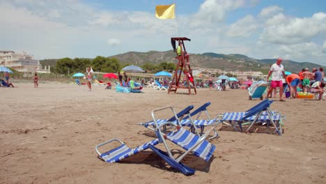 Leere-Liegestühle-Im-Sand-Mit-Gelber-Flagge,-Die-Am-Rettungsschwimmerposten-In-Alcossebre,-Costa-Del-Azahar,-Spanien,-Weht