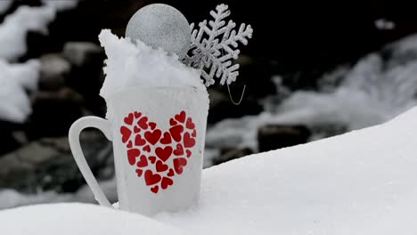 White-cup-with-a-red-hearts-design-filled-with-snow,-a-silver-Christmas-sphere-and-a-snowflake,-with-a-stream-in-the-background-in-nature