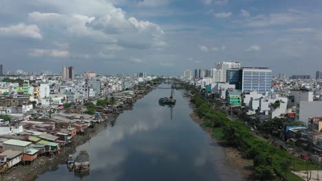 Drone-flight-along-Kenh-Te-canal-in-Ho-Chi-Minh-City-Vietnam-on-sunny-afternoon-with-river-boats,-waterfront-houses-with-traffic-bridges-and-reflections