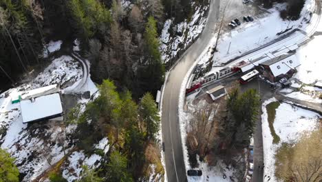 Aerial:-red-and-white-train-in-the-Alps,-emerging-from-a-tunnel-under-a-road-and-entering-a-station