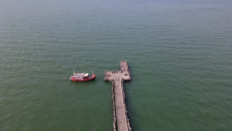 Imágenes-Aéreas-Hacia-Un-Barco-De-Pesca-Rojo-Atracado-En-El-Muelle-De-Pesca-De-Pattaya-Que-Se-Extiende-Hasta-El-Mar-En-Pattaya,-Tailandia