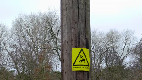 Tilt-shot-revealing-a-bright-yellow-warning-sign-mounted-on-a-wooden-gum-pole,-the-sign-indicating-the-potential-danger-and-possibility-of-death-by-the-voltage-and-current-carried-above-in-Norfolk