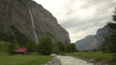 Staubbach-falls-streaming-down-steep-rocky-cliff-near-Lutschine-River-on-green-meadow-and-pine-tree-forest-on-an-overcast-day,-Lauterbrunnen,-Swiss-Alps
