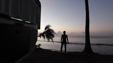 Junger-Mann-Geht-Bei-Sonnenaufgang-An-Den-Rand-Eines-Strandes-Mit-Blick-Auf-Die-Kokospalmen-Und-öffnet-Die-Tür-Des-Wohnmobils-In-Bali,-Indonesien