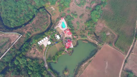 Super-high-drone-aerial-shot-in-Vadodara,-India,-over-a-house-with-a-pool-surrounded-by-a-green-forest-and-a-lake,-the-crop-fields-look-small-all-around,-like-a-digital-map