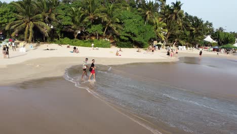 Aerial-pan-right-of-two-young-men-walking-in-sand-shore-near-sea-waves-and-palm-tree-rainforest,-Mirissa-Beach,-Sri-Lanka
