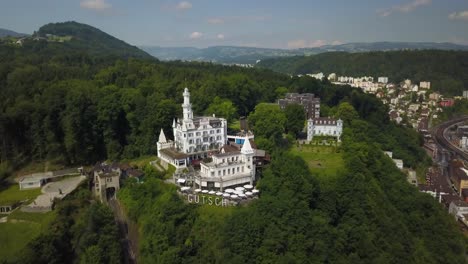 Aerial-orbit-of-Chateau-Gutsch-luxurious-hotel-on-a-hill-summit-surrounded-by-dense-forest,-near-Lucerne-Old-Town-at-daytime,-Switzerland