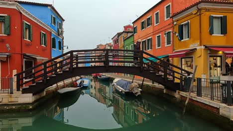 Couple-crosses-small-wooden-bridge-over-canal-of-Burano-island-in-Italy-with-colorful-houses-in-background