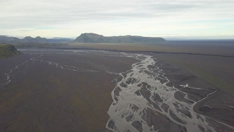 Aerial-trucking-of-stream-of-water-in-lowland-fiord-area,-verdant-hills-in-background-on-an-overcast-day,-Iceland