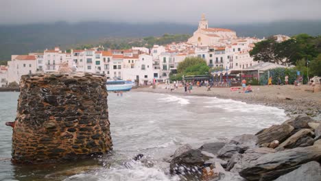 Tourists-At-The-Beach-In-Cadaques,-Spain-With-Santa-Maria-de-Cadaques-Catholic-Church-In-Background