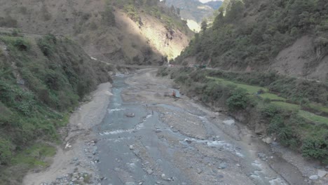Rocky-river-bed-excavator-pulling-river-rock-for-cement-production-bridge-spanning-over-water-in-valley-connecting-communities-in-Kabayan-Benguet-Philippines-reverse-reveal-ascending-aerial
