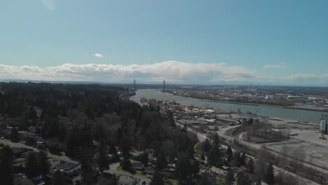 Scenic-view-of-Delta-BC-on-the-edge-of-the-Fraser-river-with-the-Alex-Fraser-bridge-in-the-background-Bright-day-blue-sky-clouds-Aerial-Wide-reversing-revealing-homes-neighbourhood-below