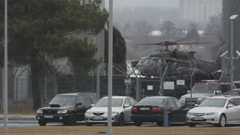 A-military-helicopter-taxis-across-an-airfield,-Civilian-cars-parked-outside-airport-security-fencing