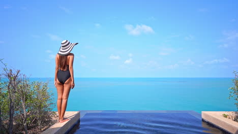 Back-View-of-Lonesome-Slender-Female-in-Swimsuit-and-Floppy-Summer-Hat-Standing-by-Infinity-Pool-With-Stunning-View-of-Tropical-Sea-Horizon