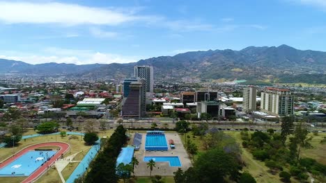 Sobrevolando-El-Parque-La-Sabana,-Dentro-De-La-Ciudad-En-San-José-En-Costa-Rica,-Mirando-Canchas-Deportivas-Y-Edificios-Altos