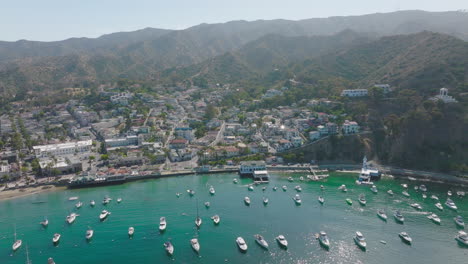 Drone-Shot-Flying-Over-Catalina-Island,-Boats-in-Harbor-with-Hotels-and-Houses-Nestled-in-Mountains
