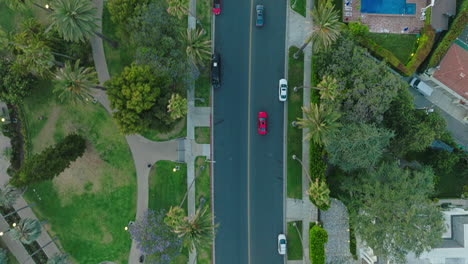 Birdseye-Drone-Shot-of-Red-and-Blue-Cars-Driving-Down-Road-in-Beverly-Hills,-Green-Grass-and-Rooftops-in-View