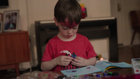 A-cute-baby-boy-with-designed-forehead-wearing-a-red-T-shirt-having-sketch-of-two-birds-opens-a-drawing-book-and-engages-in-a-work-of-art