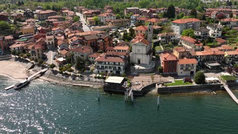 Aerial-slow-orbit-shot-of-an-Italian-village-on-the-shore-of-Lago-Maggoiore-on-a-bright-sunny-day-with-shimmering-waters,-and-classic-tile-roof-buildings