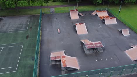 Aerial-view-flying-rising-orbit-above-fenced-skate-park-ramp-and-tennis-court-in-empty-closed-playground