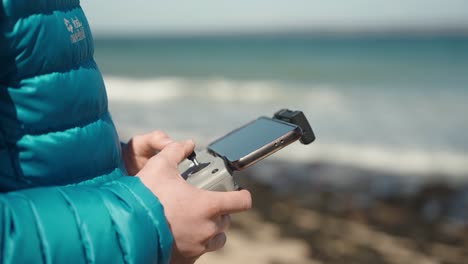 Drone-pilot-remote-controlling-flying-drone-while-wearing-a-blue-jacket-at-the-beach-on-a-sunny-day