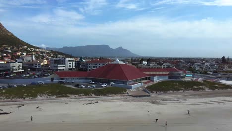 Drone-shot-of-Muizenberg-beach,-Cape-Town---drone-is-reversing-from-a-house,-revealing-the-beach-and-ocean