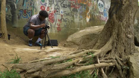 black-photographer-setting-up-camera-in-nature-in-front-of-graffitied-wall-on-a-sunny-day-by-a-tree