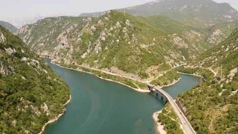 Vista-Aérea-De-Parte-Del-Río-Neretva-Con-Un-Puente-Sobre-Un-Afluente-En-Bosnia-Con-La-Carretera-Al-Pie-De-Las-Montañas