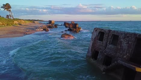 Aerial-view-of-abandoned-seaside-fortification-buildings-at-Karosta-Northern-Forts-on-the-beach-of-Baltic-sea-,-waves-splash,-golden-hour-sunset,-wide-drone-shot-moving-forward