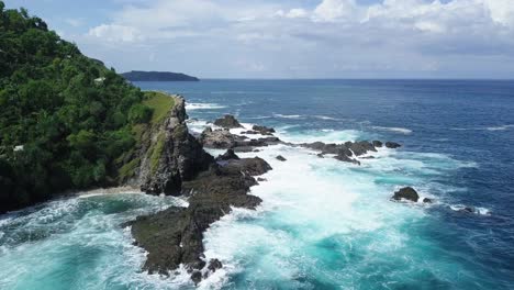 Reveal-drone-shot-of-waves-of-Indian-Ocean-hitting-boulder-and-hill-overgrown-by-dense-of-trees-beside-the-beach-in-sunny-condition-weather---Pengilon-Hill,-Indonesia,-Asia