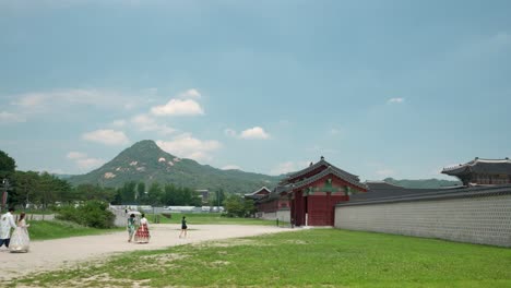 A-couple-in-Hanbok-attire-and-other-tourists-enter-Yongseongmun-Gate-to-Gyeongbokgung-Palace-with-Bukhansan-mount-summit-against-cloudy-blue-sky---wide-landscape