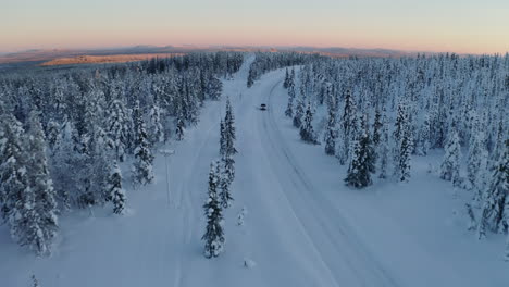 Aerial-view-above-snowy-Scandinavia-landscape-following-vehicle-on-journey-along-long-curved-remote-road-through-Lapland-woodland