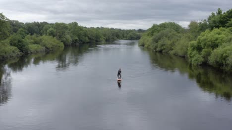 Standup-paddleboarder-paddles-toward-camera-on-green-country-river