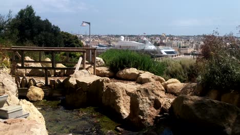 Peaceful-river-fountain-in-Herbert-Ganado-park-in-Floriana,-Malta-with-large-cruise-ship-and-the-three-cities-in-background