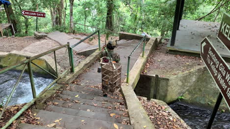 Balinesischer-Einheimischer-Geht-Stufen-über-Eine-Kleine-Brücke-Hinunter-Und-Trägt-Einen-Vogelkäfig-Mit-Bambusstock