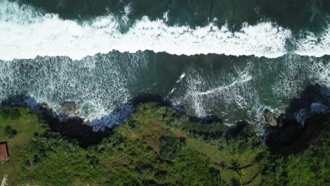 Aerial-top-down-of-massive-waves-of-sea-crashing-against-overgrown-cliffs-during-sunlight